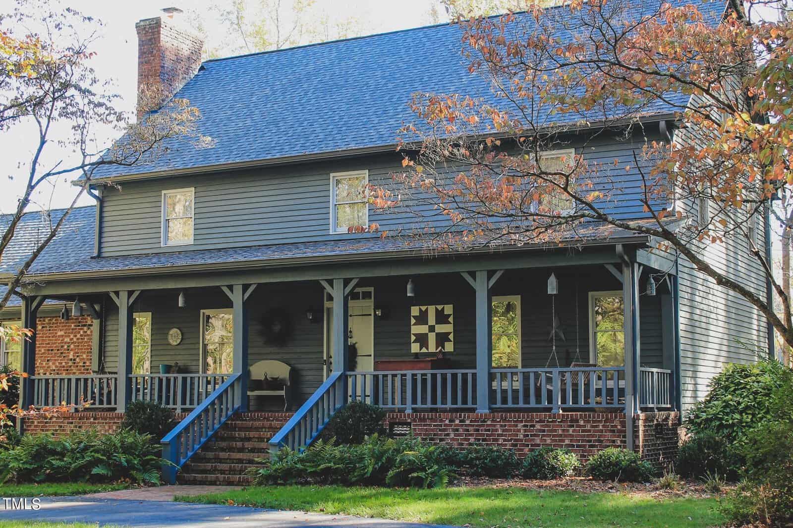 A two-story house with a dark blue exterior, white trim, a covered front porch with blue stairs, and a brick foundation surrounded by mature trees with autumn foliage.