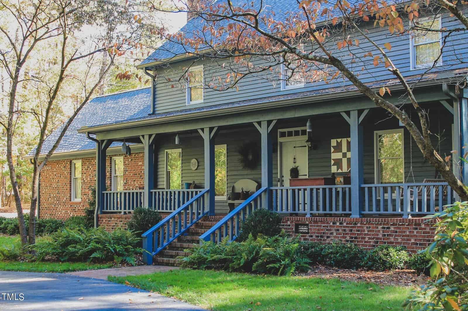A two-story house with a brick exterior and a wooden porch featuring blue steps, surrounded by trees with autumn foliage.