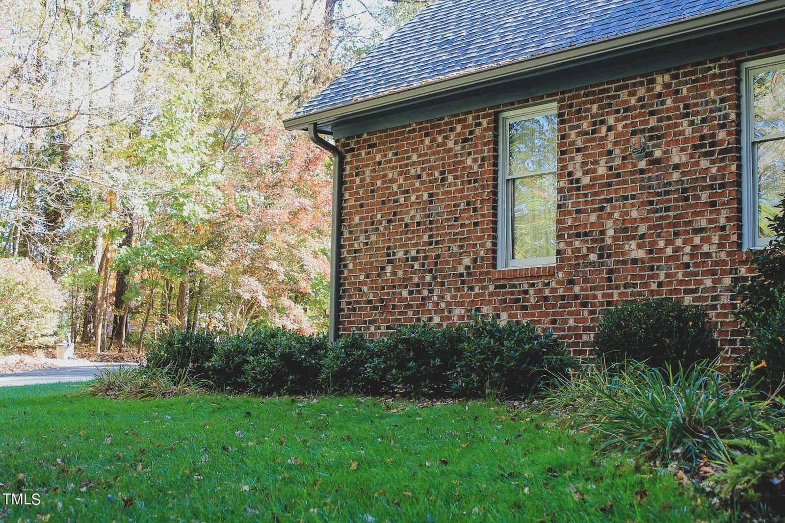 A brick house with a single window is surrounded by greenery and a lawn, with a pathway leading away from the house.