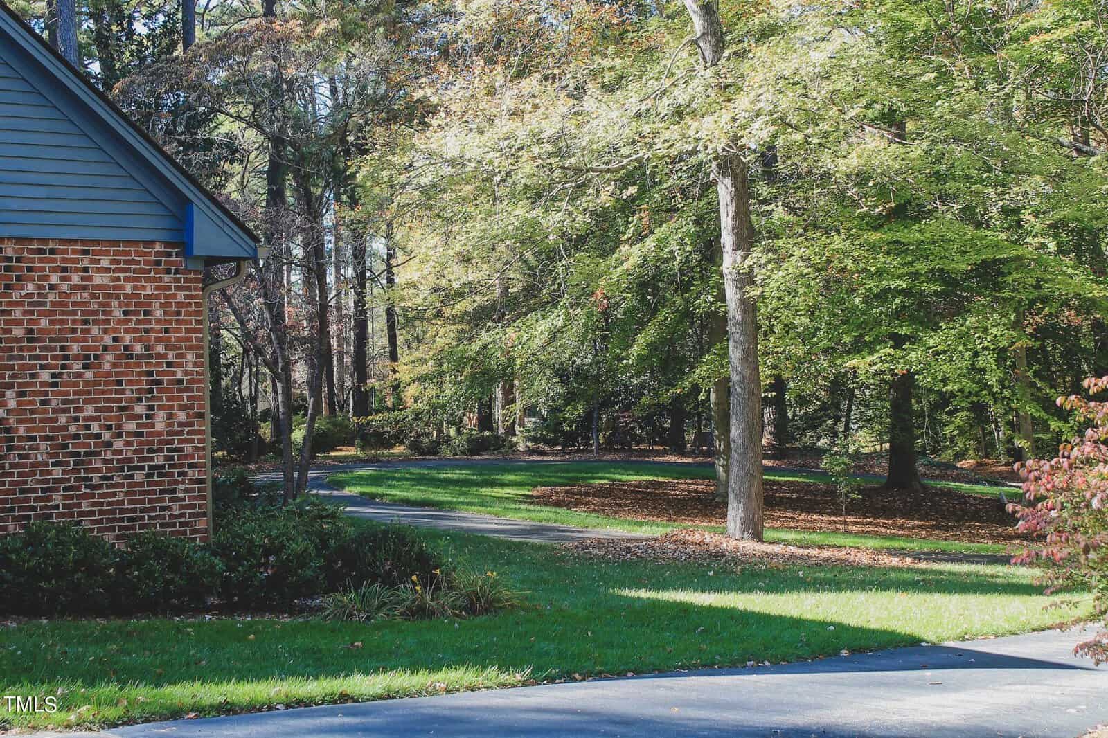 A winding path leads through a serene, tree-lined landscape on a sunny day, with the corner of a brick building visible to the left.
