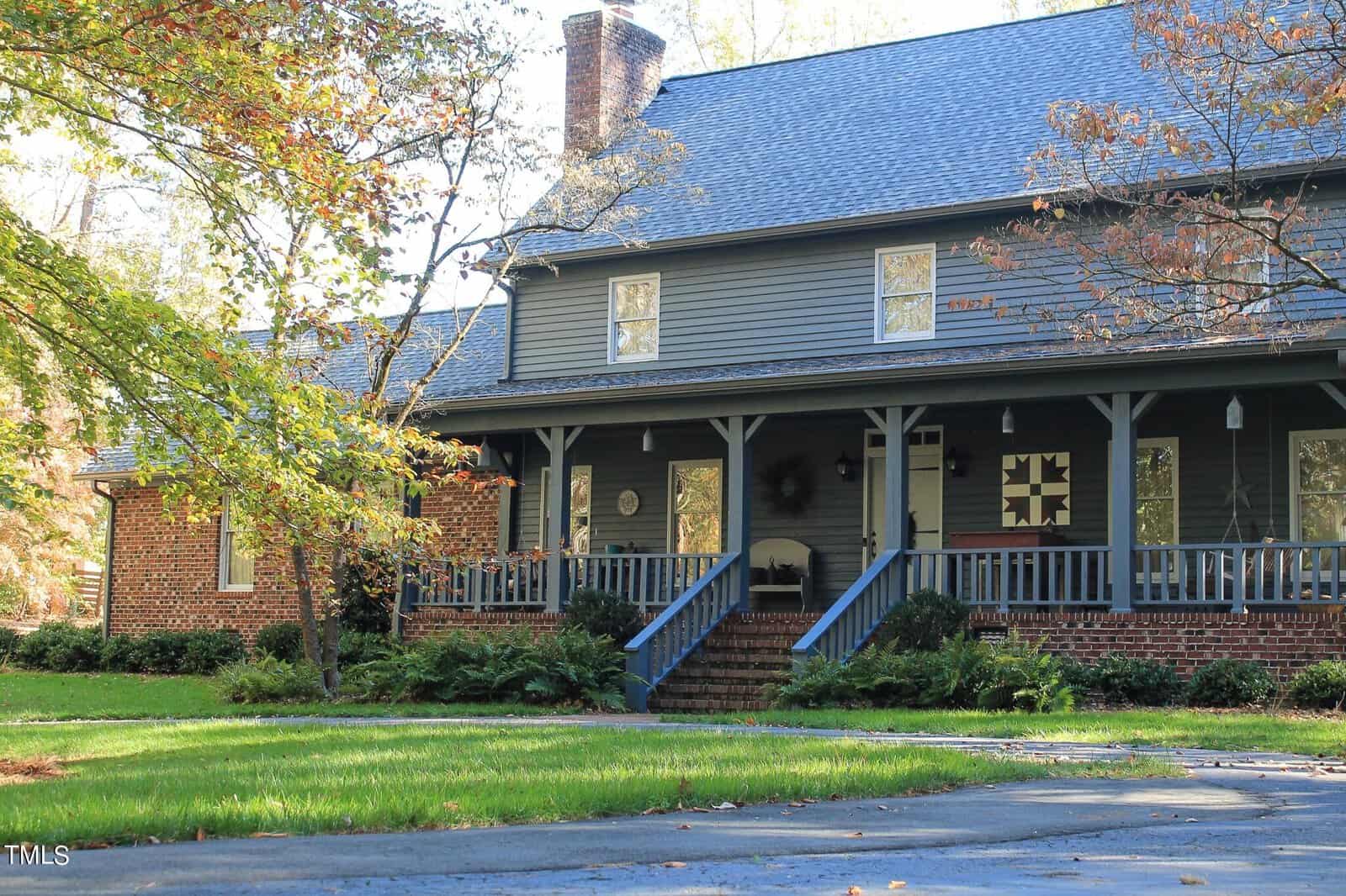 A traditional two-story house with a brick foundation and a large covered front porch surrounded by mature trees.