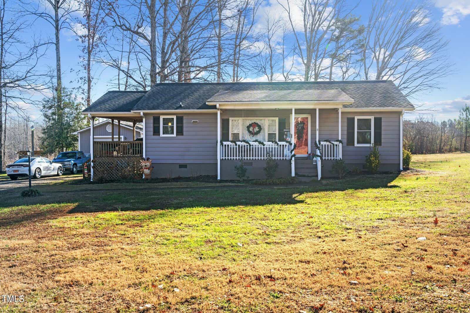 A small gray house at 3202 Little Mountain Creek Road, Oxford, boasts a front porch decorated for the holidays with a wreath and red ribbon on the door. The brown lawn, framed by trees, sits under a clear sunny sky, while a driveway and additional structure are visible to the left.