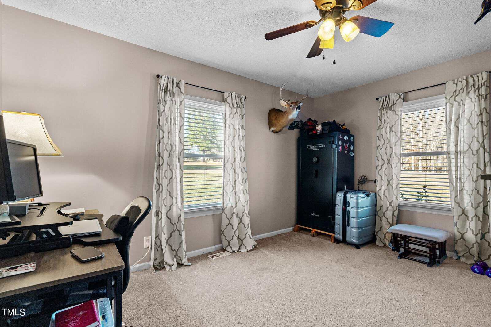 A home office with a desk, computer, and chair. A ceiling fan is on above. Two windows with patterned curtains let in natural light. A safe and a mounted deer head are on one wall. A small stool and weights are on the carpeted floor.