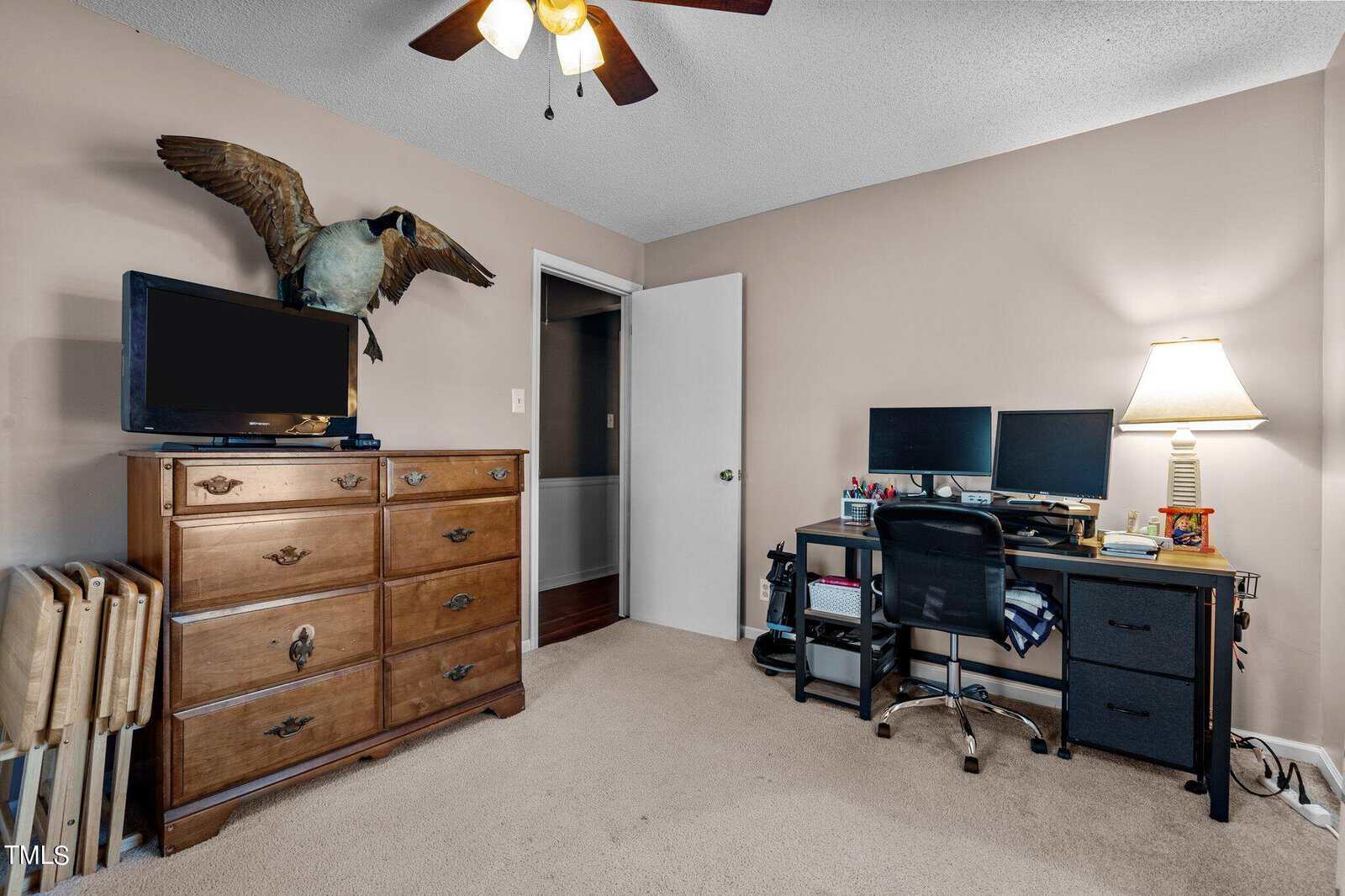A cozy home office with a desk featuring two monitors and various supplies. A lamp adds light to the room. A dresser with a TV and a large decorative bird sculpture sits opposite the desk. The door is slightly open, revealing a hallway.