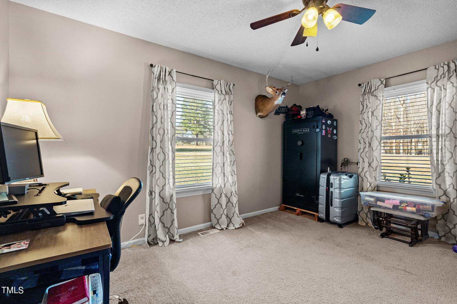 A home office with beige walls and a carpeted floor. It has a desk with a computer, a chair, and a lamp. Two windows with patterned curtains. A black safe and a mounted deer head are also visible in the room.
