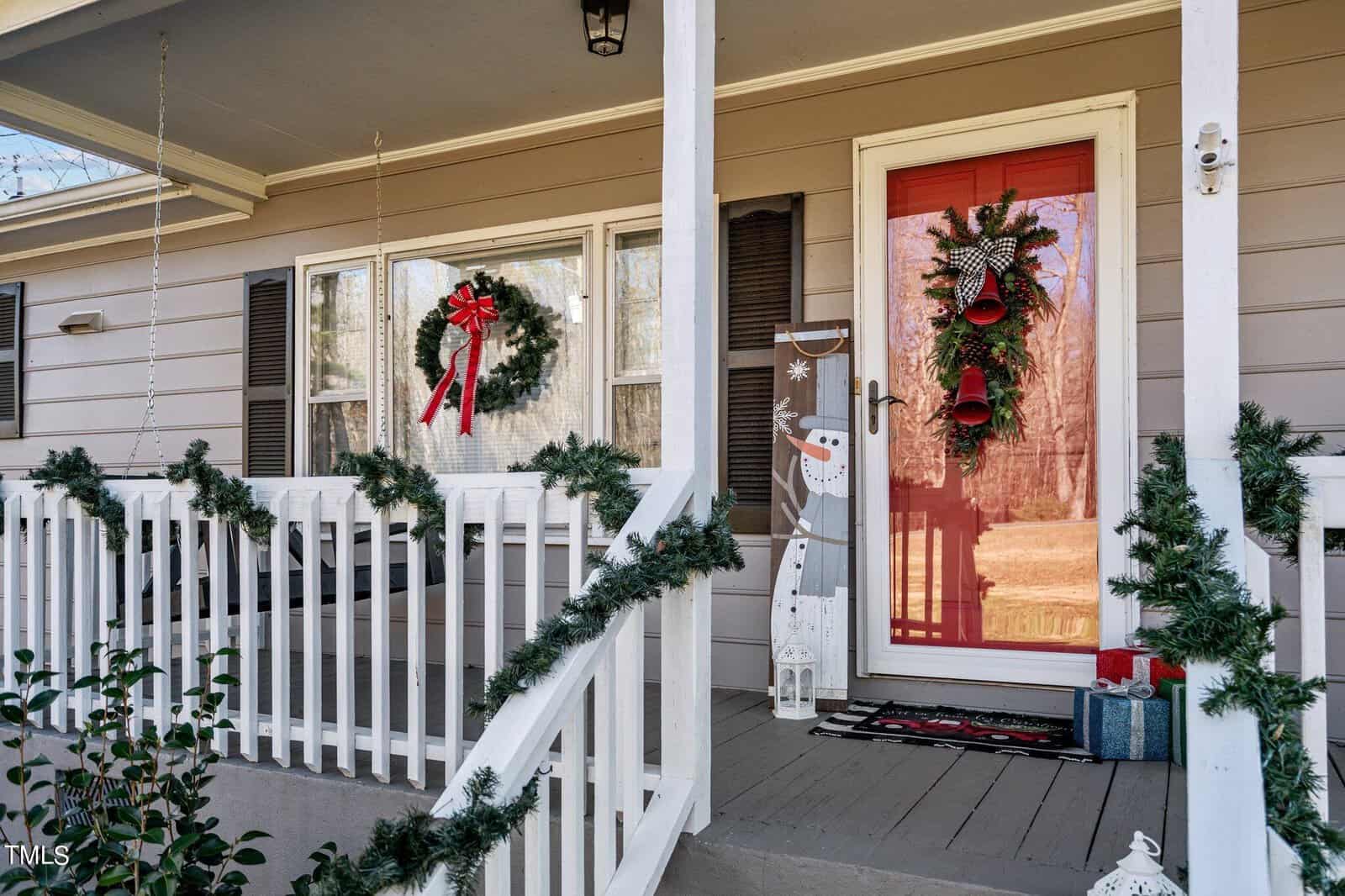 A cozy front porch decorated for the holidays with a wreath and red bow on the window, garland on the railing, a snowman figure near the door, and a red door adorned with festive greenery and bells. A welcome mat and gift boxes complete the scene.