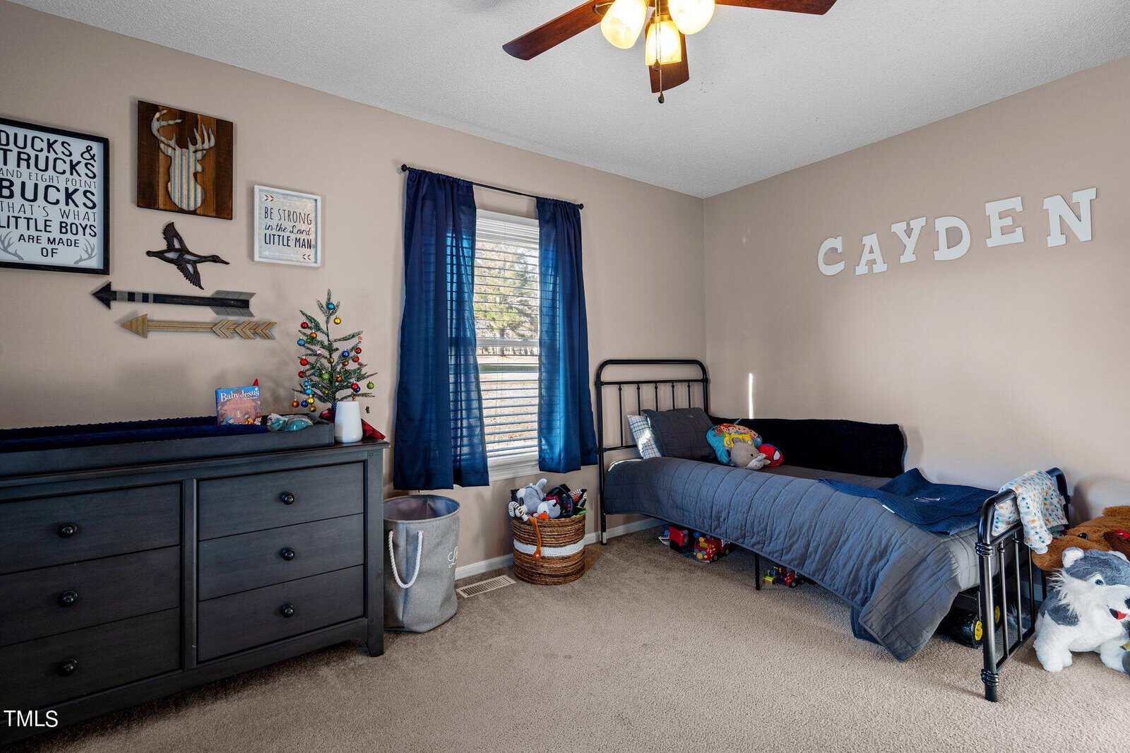 A child's bedroom with a bed, blue blanket, and toys. The walls have decorations, including a reindeer head, arrow, and wooden sign. The name "CAYDEN" is displayed in large letters. A dresser with a small Christmas tree stands in the room.