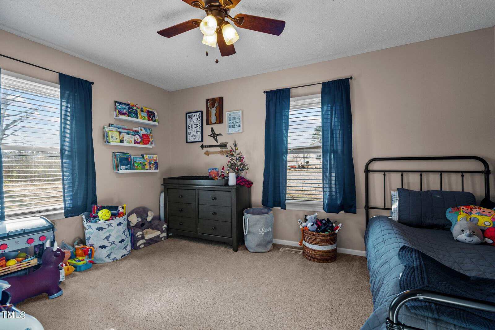A cozy child's bedroom with beige walls, a ceiling fan, and two windows with blue curtains. A bed with dark bedding is on the right, shelves filled with books are on the left, and toys are scattered around the room. A dresser is against the wall.