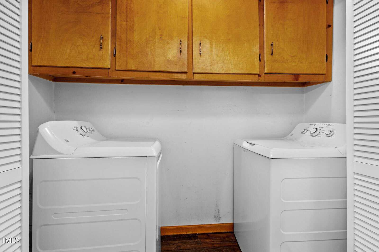 A laundry room with a white washer and dryer side by side. Above them are wooden cabinets for storage. The walls are plain white, and the floor is made of dark wood.