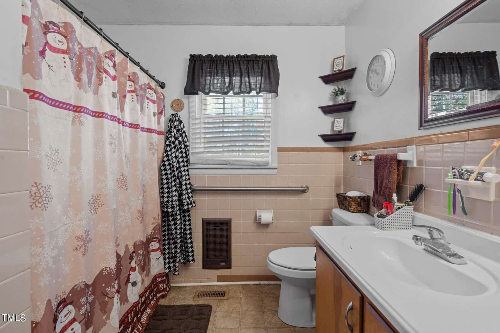 A bathroom with a snowman-themed shower curtain, a white sink with toiletries, and a toilet. A window with a dark valance, a towel rack, and shelves with decor are also visible. The walls are tiled in beige.