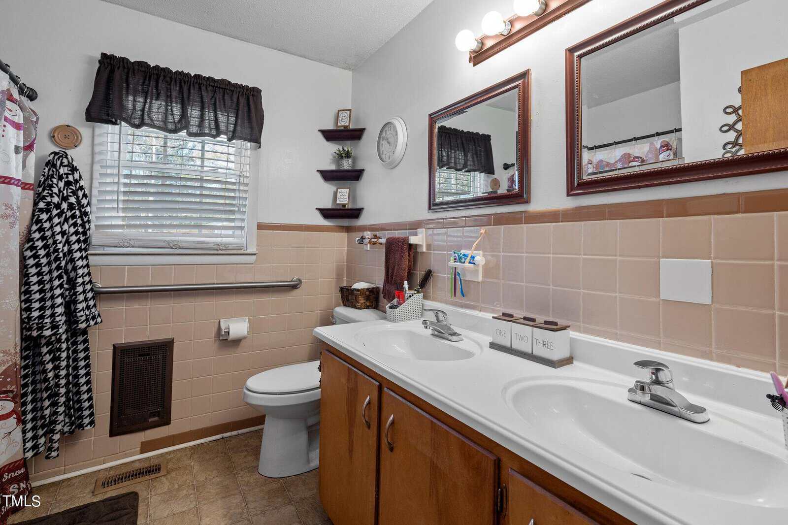 A bathroom with a double sink vanity, featuring a large mirror and two lights above. There's a toilet next to a window with a black curtain, tan tiles on the walls, and a patterned shower curtain. Various toiletries are placed around the sink area.