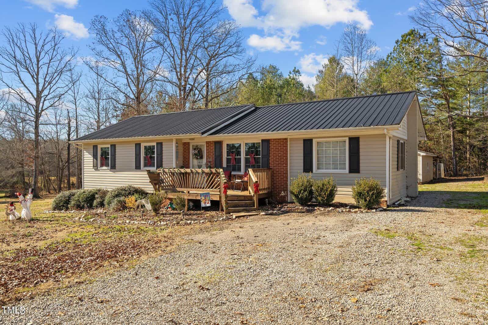 A single-story house with a beige exterior, black roof, and a front porch decorated with red accents. The lawn is bordered by leafless trees and a gravel driveway, set against a clear blue sky.
