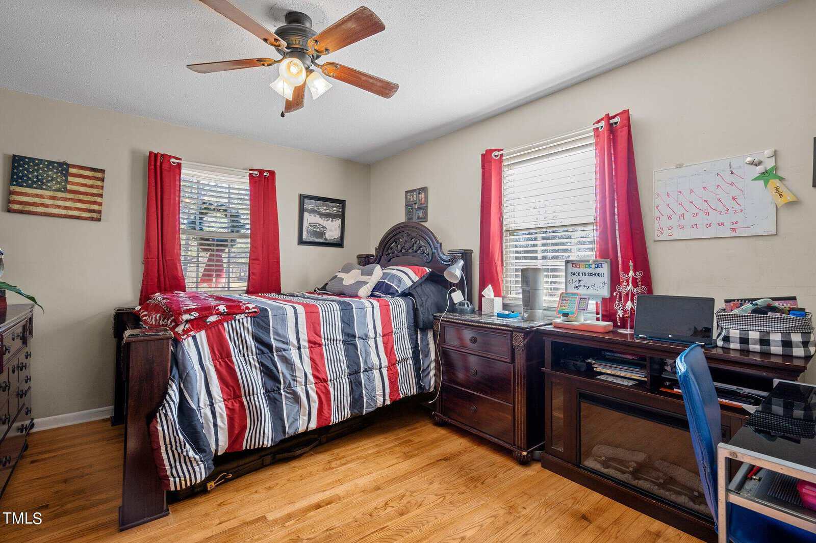 A small bedroom with a twin bed featuring a red, white, and blue striped comforter. The room has wooden furniture, red curtains, a ceiling fan, and two windows. A desk with office supplies is on one side, and a U.S. flag is on the wall.