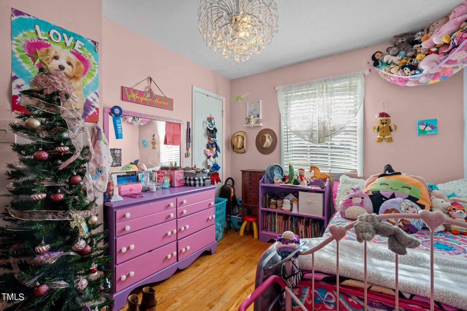 A colorful children's bedroom adorned with stuffed animals, a small Christmas tree, and vibrant pink furniture. A whimsical chandelier hangs from the ceiling, and a bed is decorated with plush toys and pillows. Sunlight streams through the window.