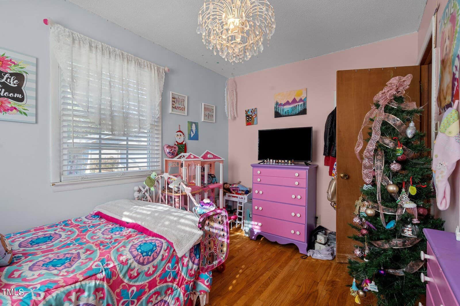 A colorful children's bedroom with a pink and purple theme. There's a bed with patterned bedding, a dollhouse, a dresser with a TV, and a decorated Christmas tree. A chandelier hangs from the ceiling, and sunlight streams through the window.