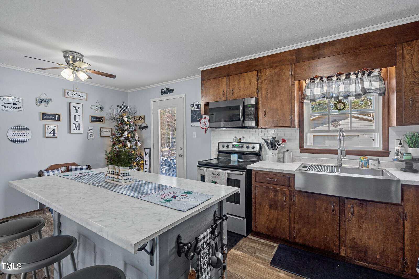 A cozy kitchen with wooden cabinets and a marble island with bar stools. The stove and sink area are clean and organized. A small decorated Christmas tree sits on the island. Light streams in through a window above the sink, creating a warm atmosphere.
