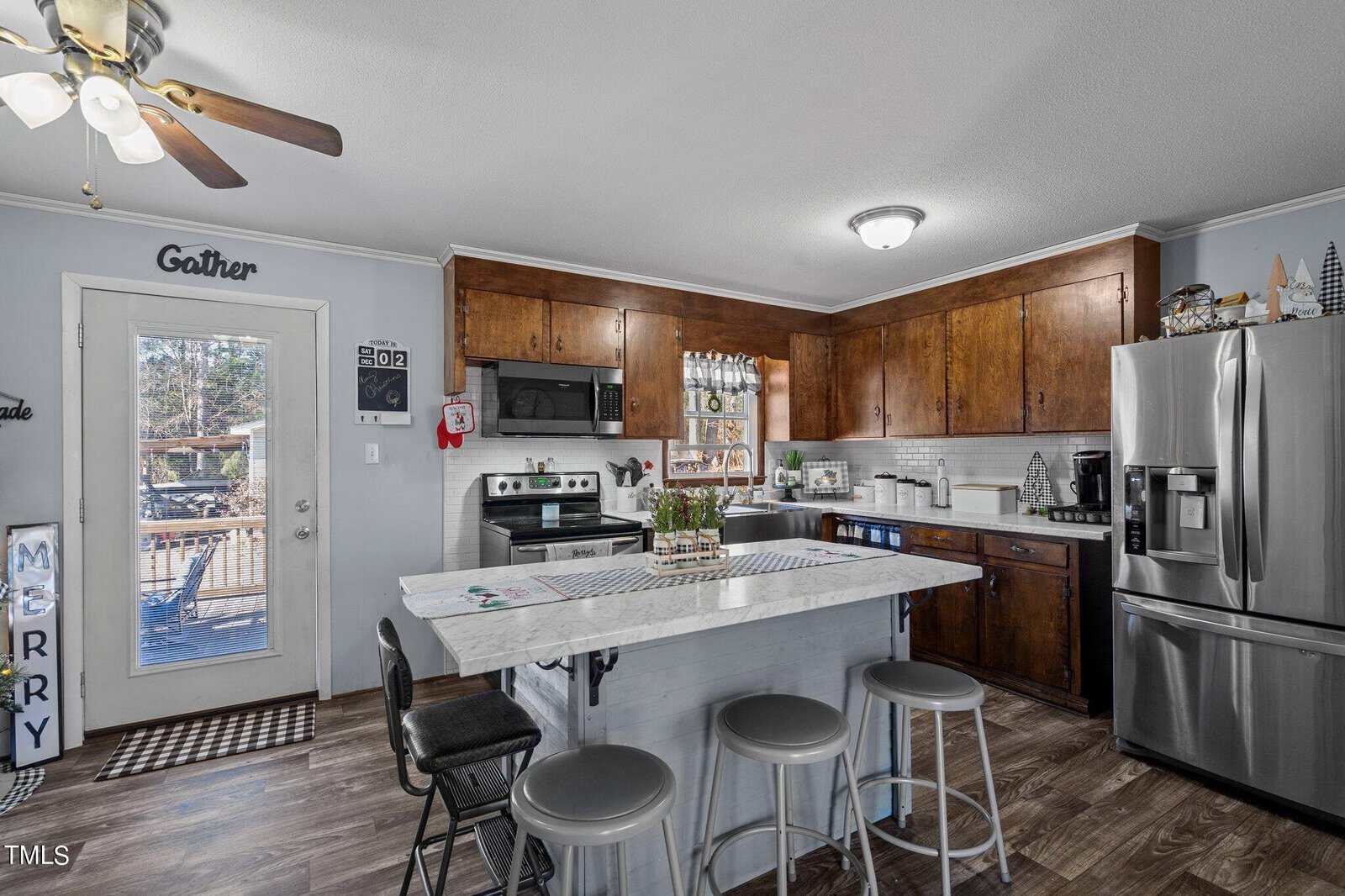 A cozy kitchen with wooden cabinets, marble island with stools, and stainless steel appliances. Seasonal decorations include letters spelling "MERRY" and various small ornaments. A ceiling fan is visible above.