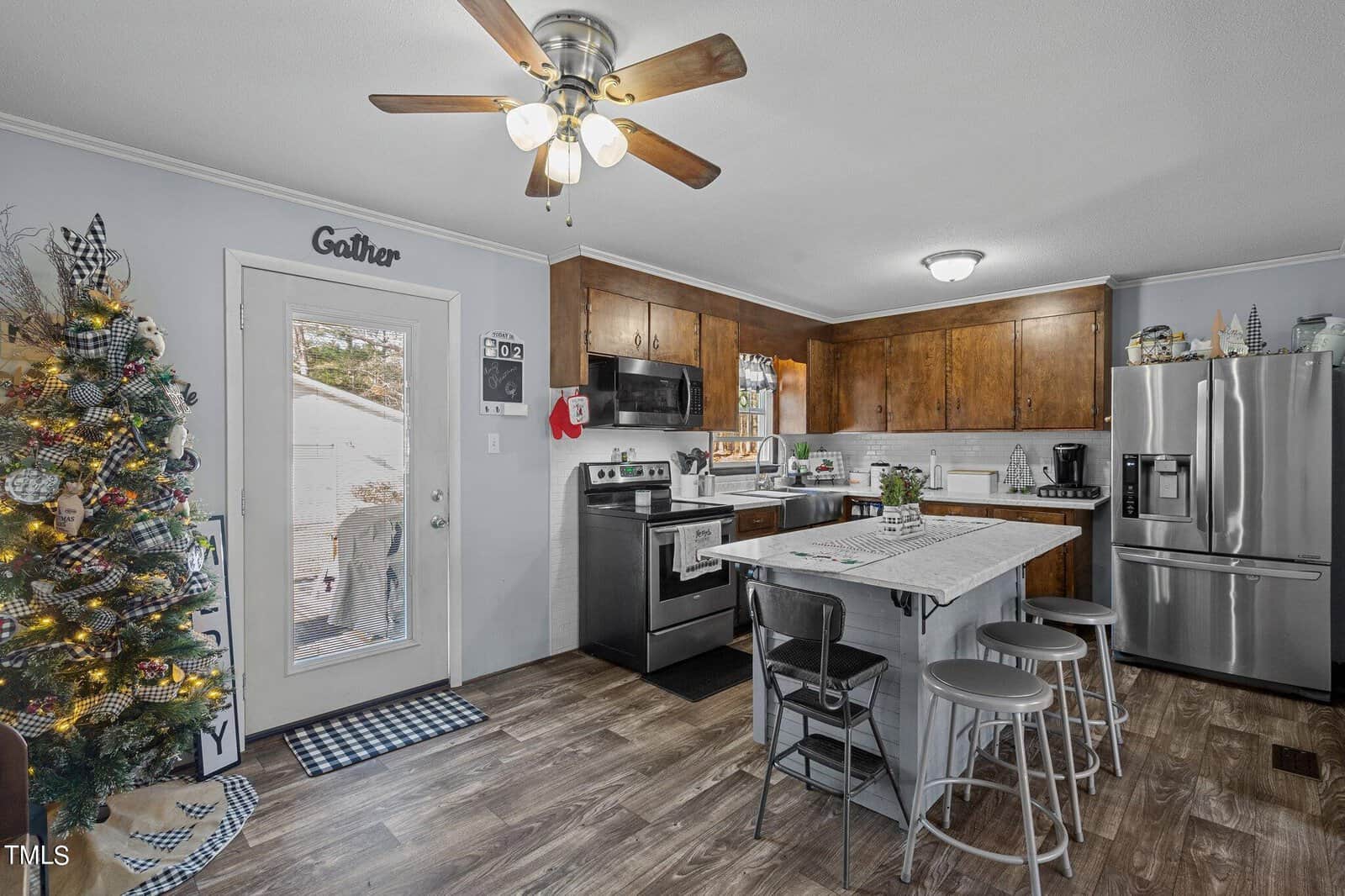A modern kitchen with wooden cabinets, stainless steel appliances, and a central island with bar stools. A decorated Christmas tree stands by the door, enhancing the festive atmosphere. The room has a ceiling fan and wood-style flooring.