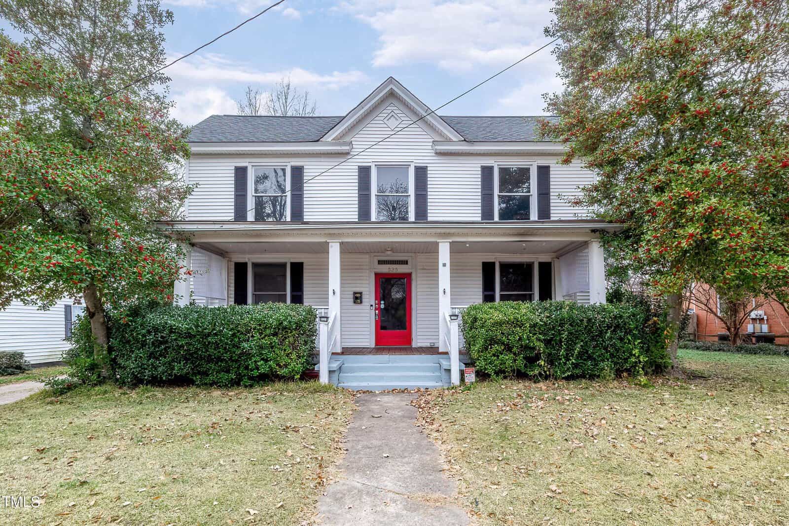A two-story white house with a red front door, located at 535 Rowland Street, Henderson NC, is surrounded by greenery and a lawn in the front.