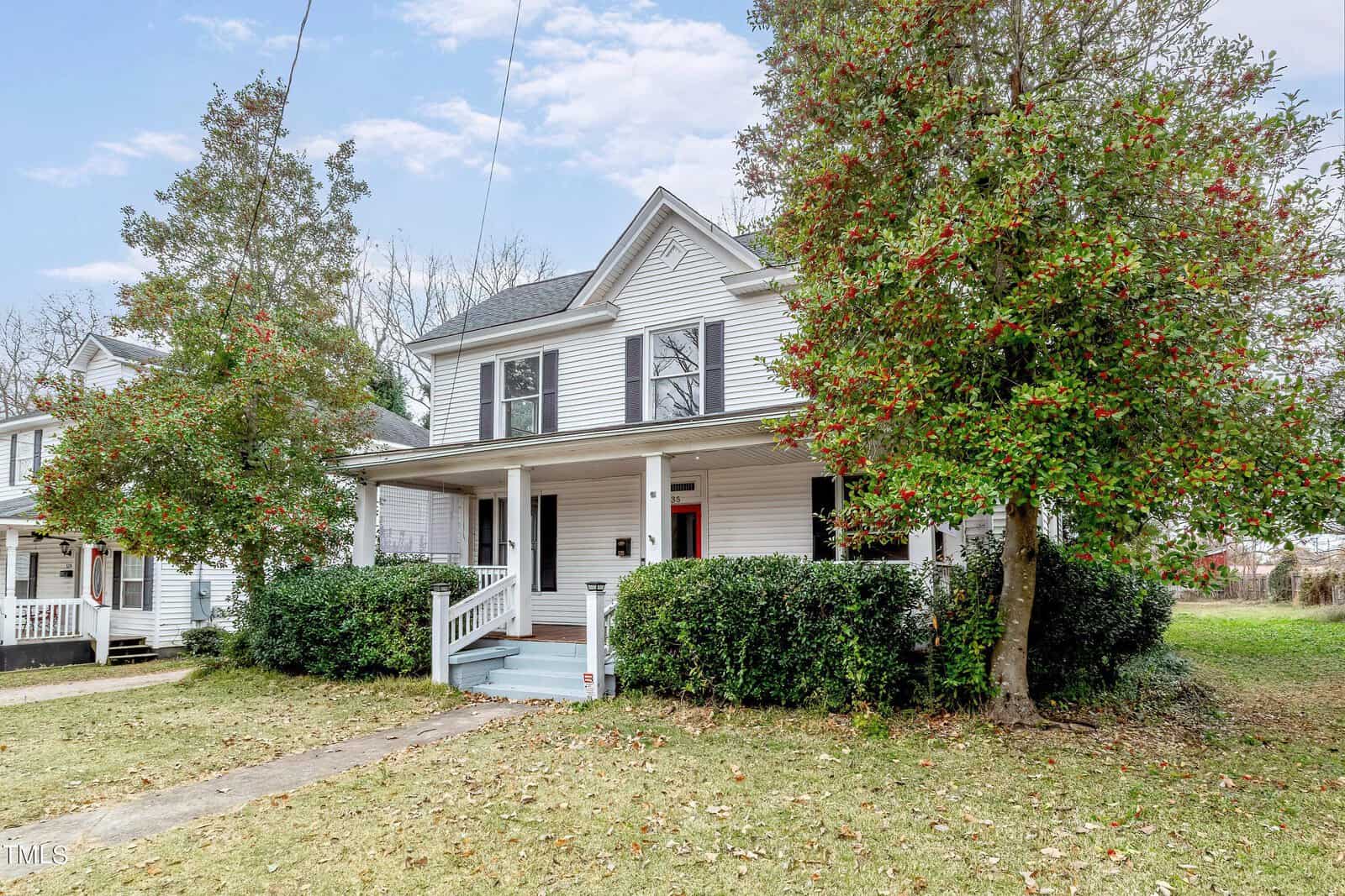 Traditional two-story white house with red door and front porch, flanked by a green tree with red berries and a partially visible neighboring house.