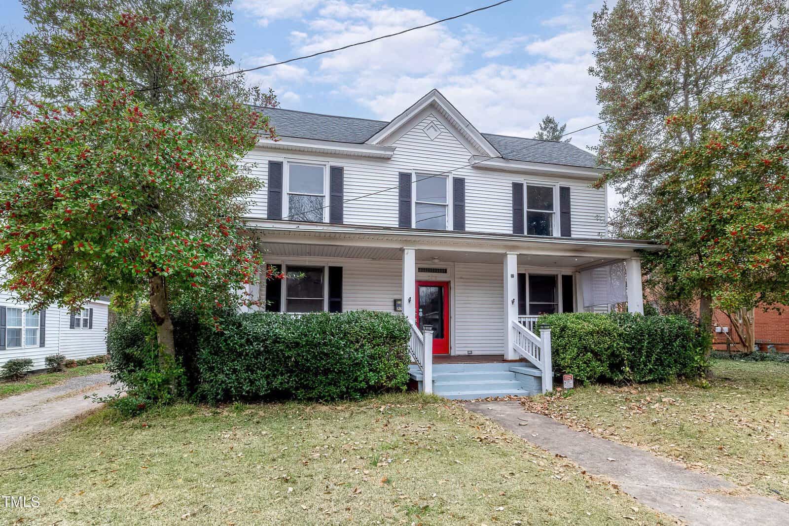 A two-story white house with a covered front porch, red door, and surrounded by greenery and trees.