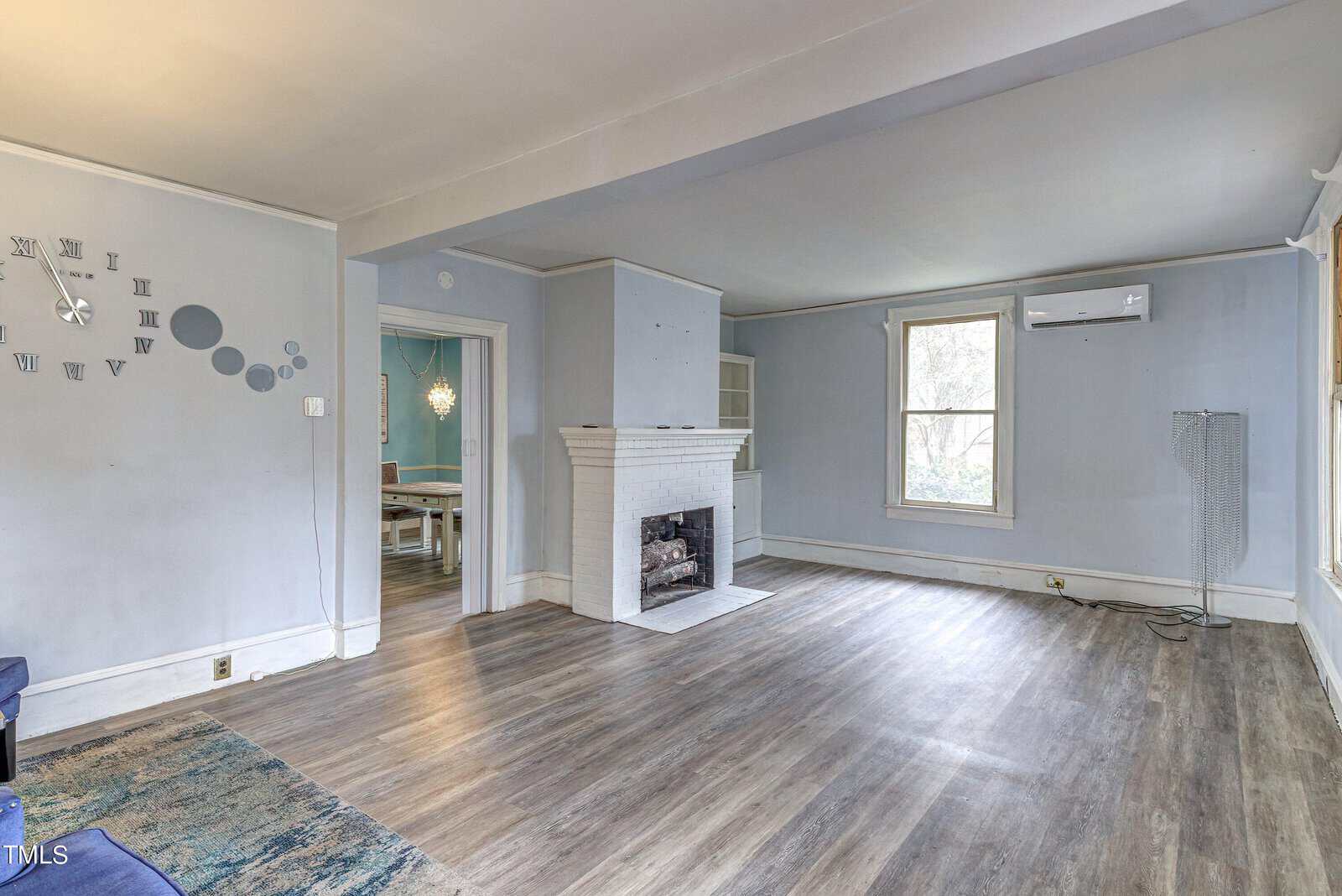 Empty room with a gray laminate floor, white walls, a fireplace with a white mantel, decorative wall clock accents, and a standalone fan to the left side.