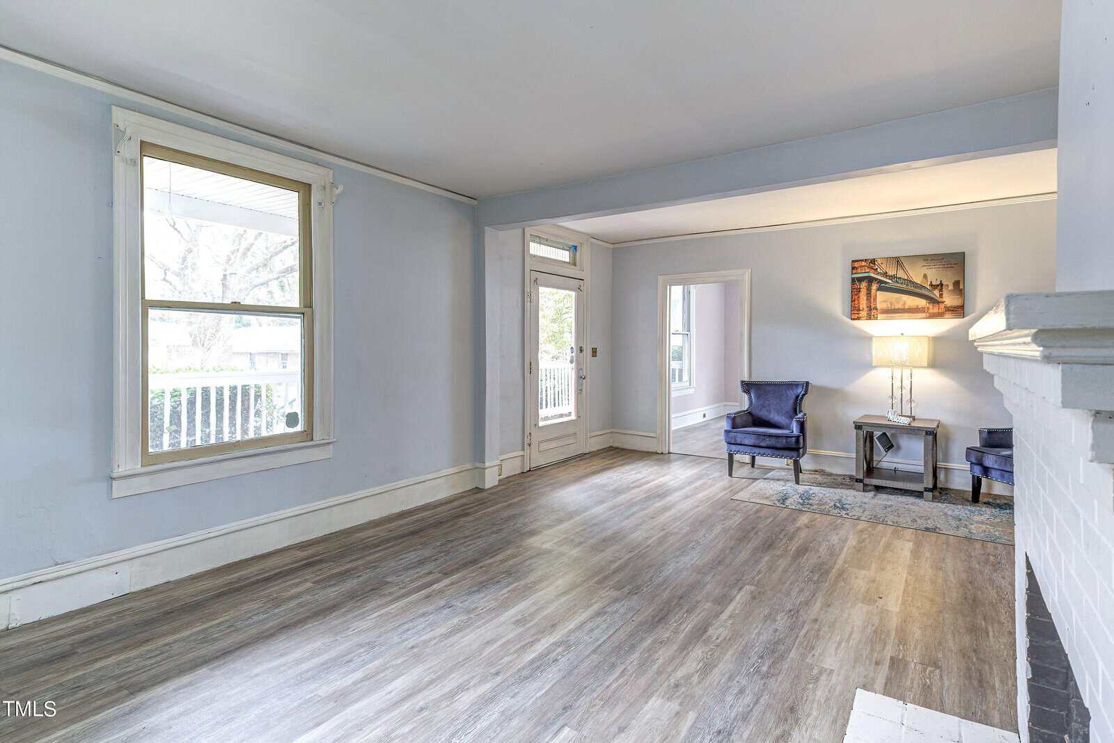 An empty living room with hardwood floors, soft blue walls, and a white trim. a fireplace is on the right, flanked by a chair and a lamp, with a painting above it. two windows provide natural light, and the front door is visible in the background.