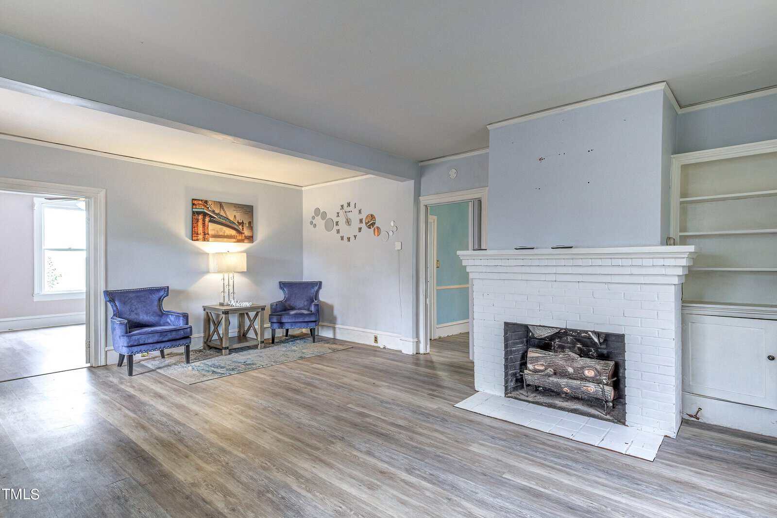 A spacious room featuring a white fireplace, two blue armchairs, and built-in shelves, with light gray walls and laminate flooring.