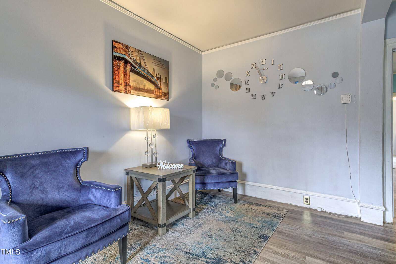 A well-lit room with two blue accent chairs, a side table with a lamp, a decorative rug, and a wall-mounted clock design with scattered numbers.