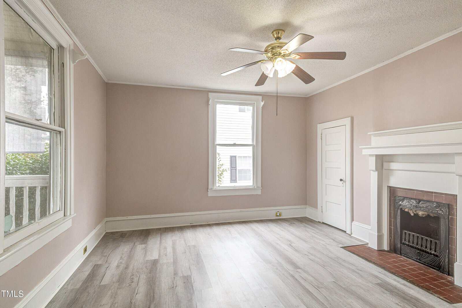 A vacant room with light mauve walls, a white ceiling fan, two windows providing natural light, a white fireplace, and laminate flooring.
