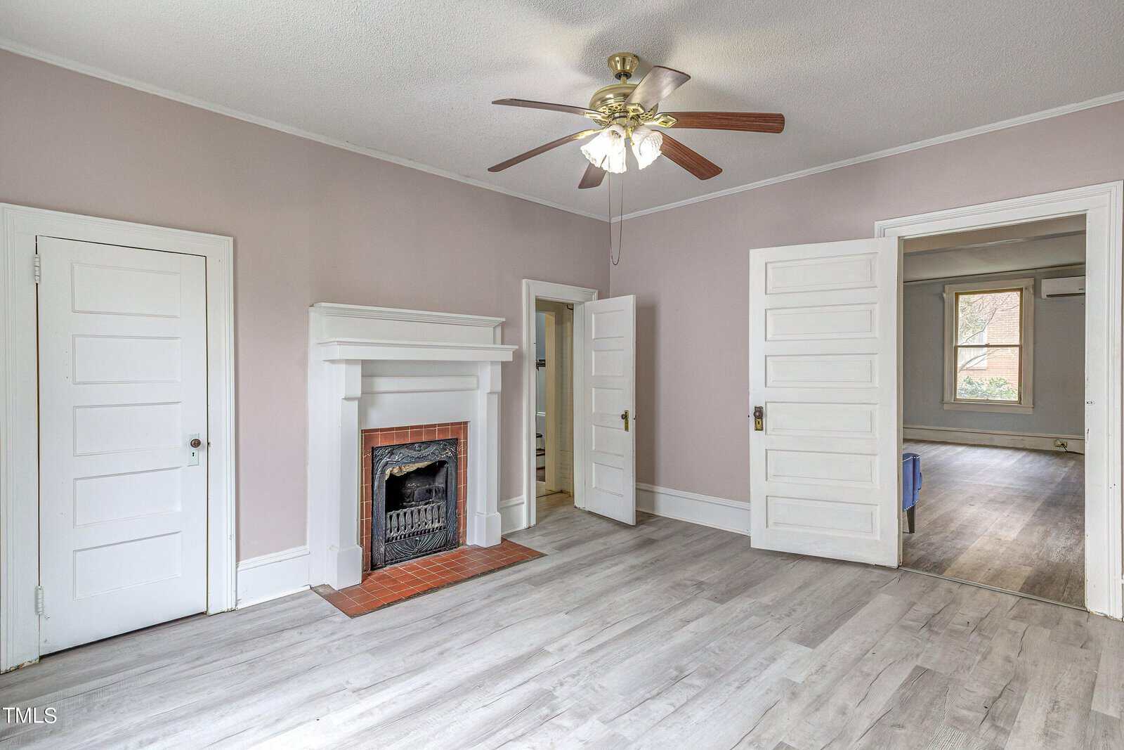 An empty room featuring a light-gray wood flooring, a pastel-pink wall, a white fireplace with a brick interior, ceiling fan with lights, and two open doorways.