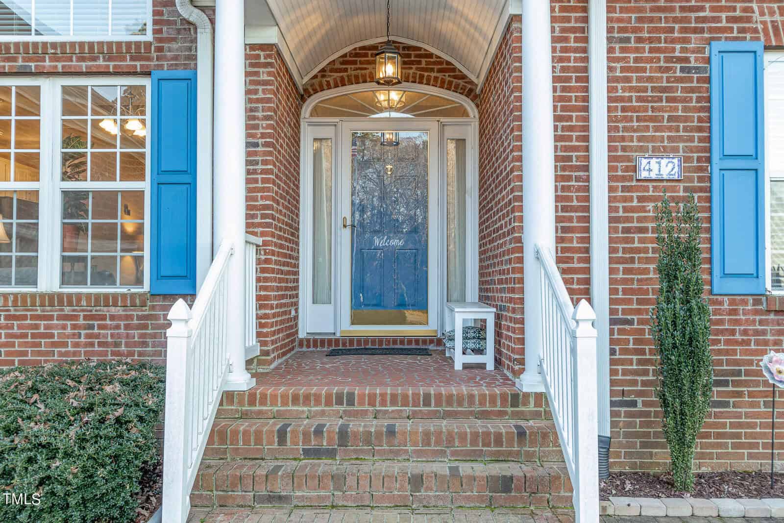 Front entrance of a brick house with white columns and blue shutters. The door has a glass pane with "Welcome" written on it. Steps lead up to the porch, adorned with a small table and plant. The house number "412" is displayed on the wall.