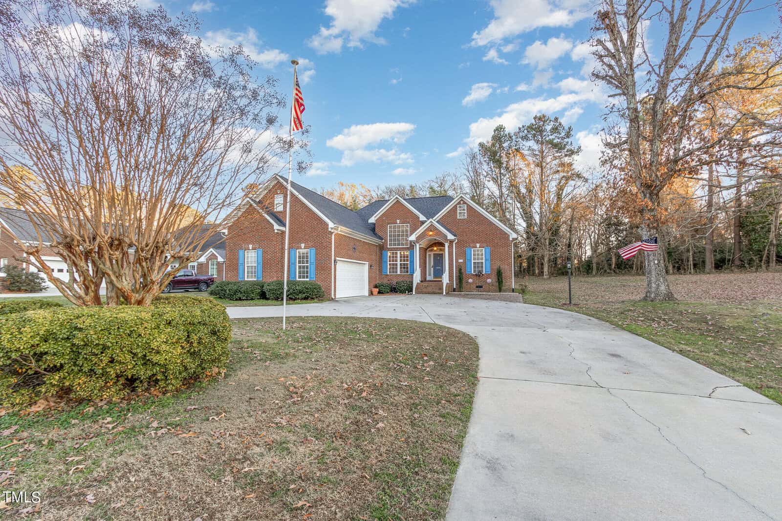 A brick house at 412 E Spring Street, Oxford, featuring a double garage, welcomes visitors at the end of a curved driveway. An American flag waves proudly in the front yard. In the background, trees with autumn leaves create a vibrant tapestry under a partly cloudy sky.
