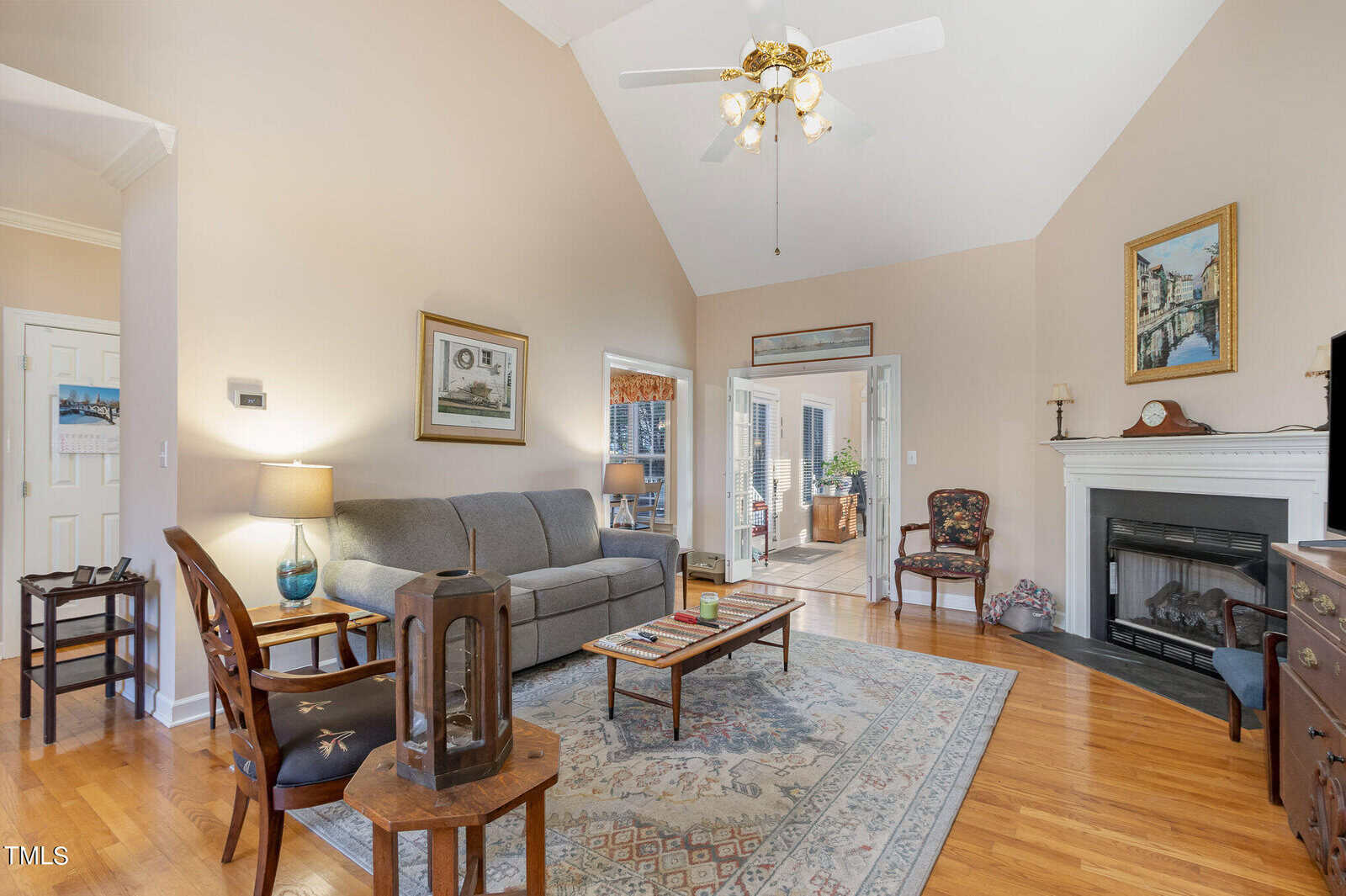 A cozy living room with a high ceiling features a gray sofa, a wooden coffee table, and an armchair. There's a fireplace on the right, and a ceiling fan above. The room has hardwood floors and is decorated with framed artwork and a patterned rug.