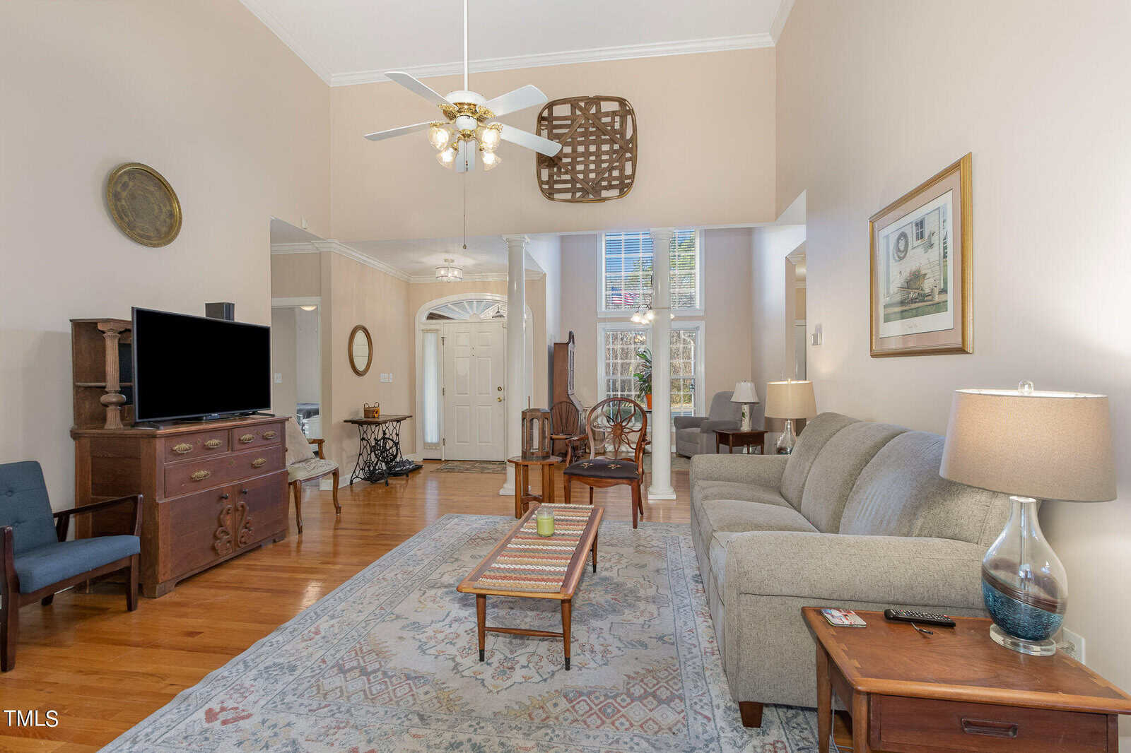 Spacious living room with high ceilings, featuring a gray couch, a wooden coffee table, and a TV on a wooden stand. A decorative rug covers the hardwood floor. Large windows and a light fixture with ceiling fans provide ample light.