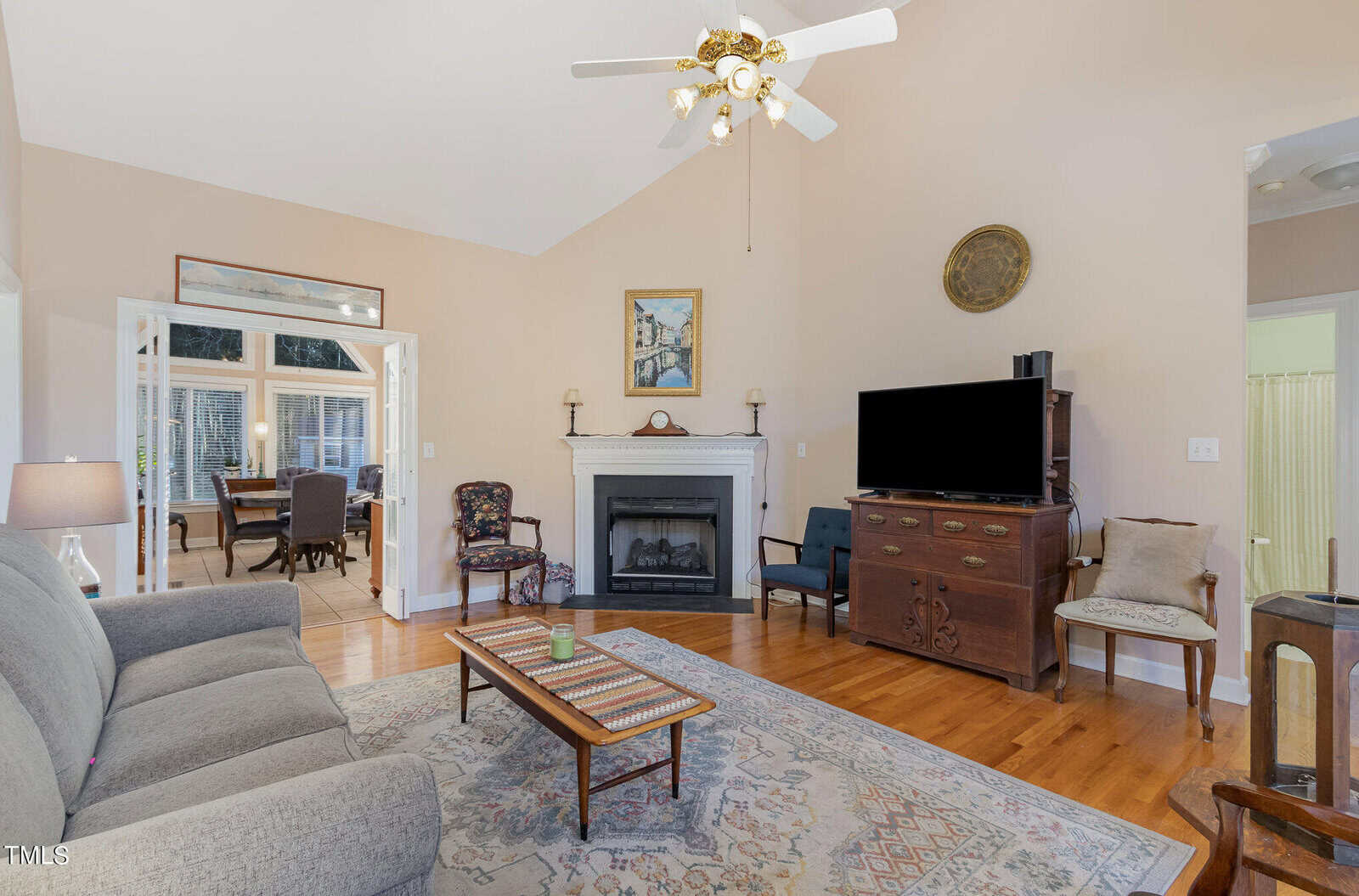 A cozy living room with a gray sofa, colorful area rug, and wooden flooring. A fireplace is centered under a framed painting, and a ceiling fan light fixture is above. A flat-screen TV sits on a wooden cabinet near an armchair.