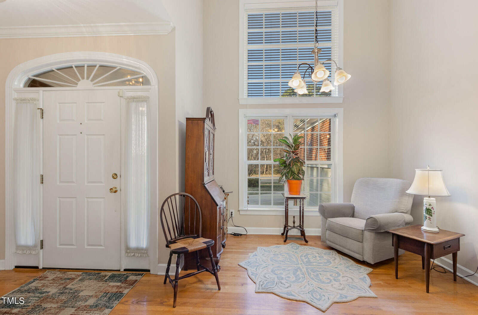A bright living room with a white door and arched window. There's a high ceiling with a chandelier, an armchair next to a small wooden table with a lamp, and a large potted plant by a window. A wooden chair and cabinet sit nearby.