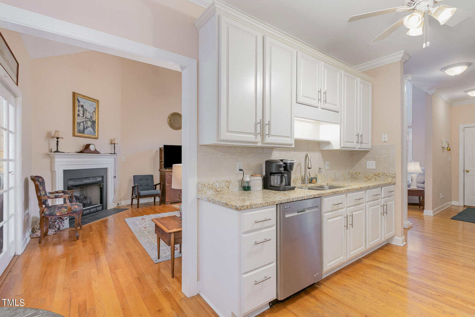 Bright kitchen with white cabinets, granite countertops, and stainless steel appliances, including a dishwasher and coffee maker. The room opens to a living area with hardwood floors and a fireplace. A ceiling fan is above, and a hallway is visible to the right.