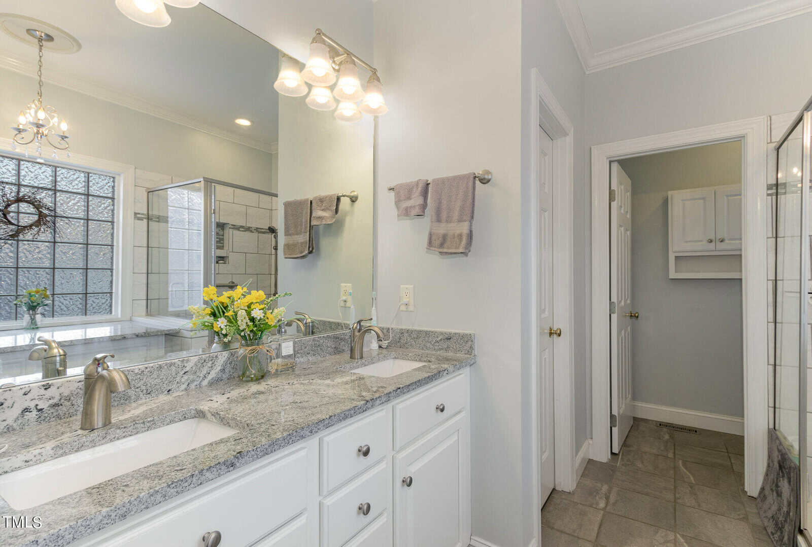A bright bathroom with a dual-sink vanity featuring a marble countertop and chrome faucets. A mirror runs the length of the vanity. Yellow flowers in a vase and towels are on the counter. The room includes a glass shower enclosure and a window.