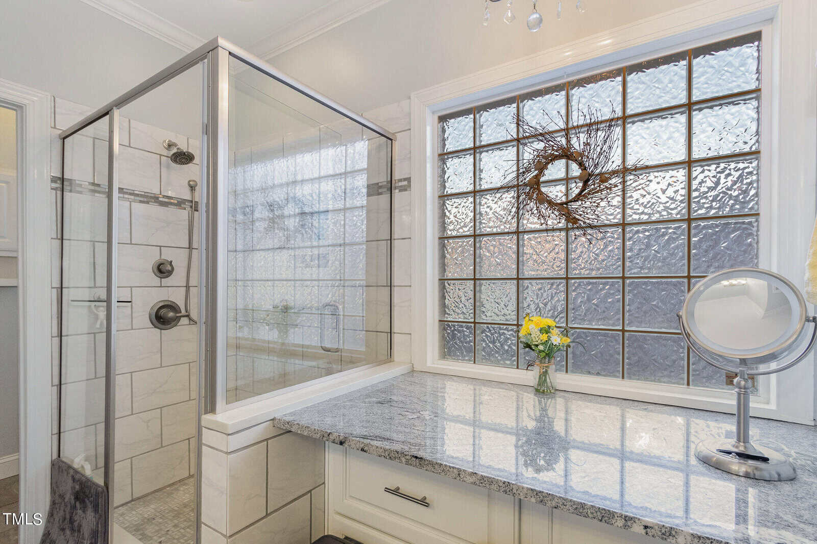 A bathroom with a glass-enclosed shower, featuring white tiles and a sleek design. A granite countertop with a vase of yellow flowers and a round mirror is next to a patterned glass window adorned with a decorative wreath.