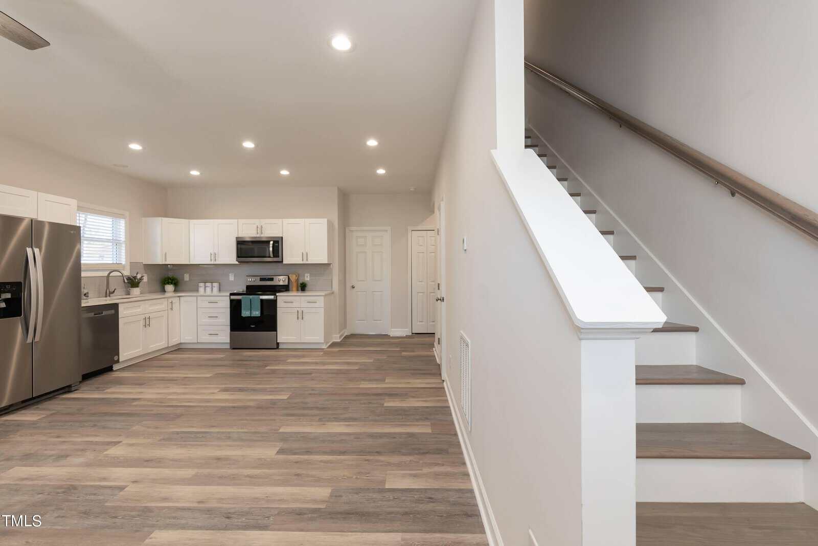 A modern kitchen with stainless steel appliances, white cabinetry, and wood-style flooring. To the right, a staircase with a wooden handrail leads upstairs. The room is well-lit with recessed ceiling lights.