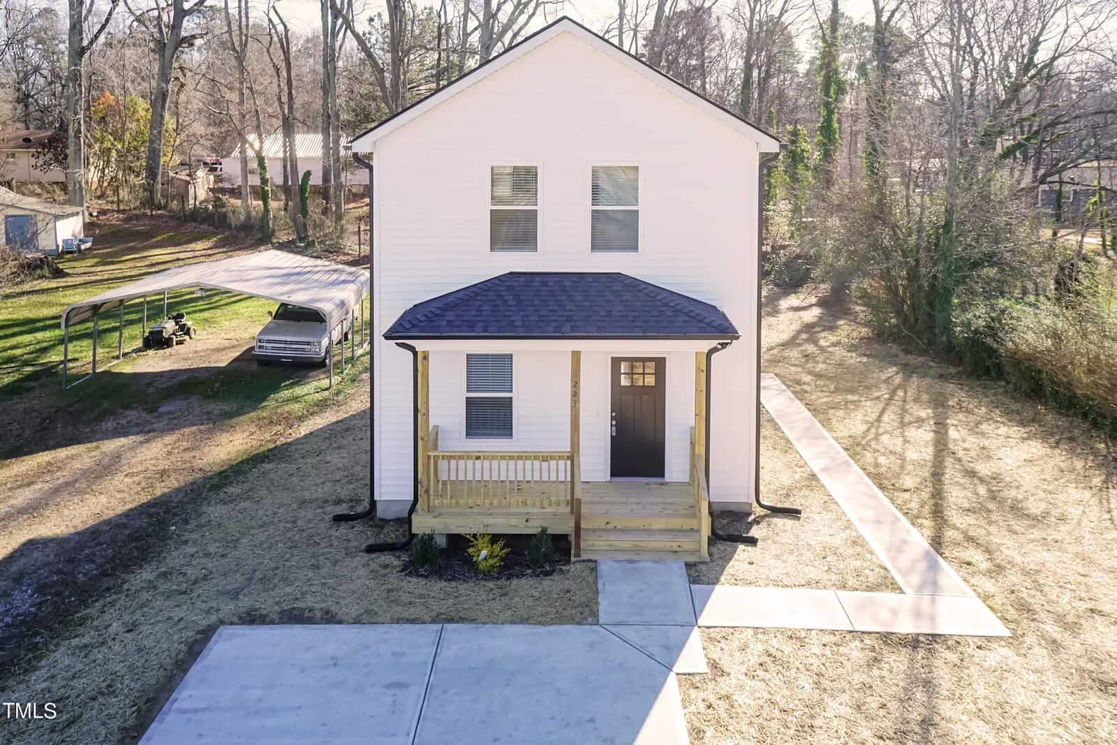 A two-story white house with a dark roof, small wooden porch, and concrete driveway. It's situated on a grassy lot surrounded by leafless trees. A metal carport is visible on the left side.
