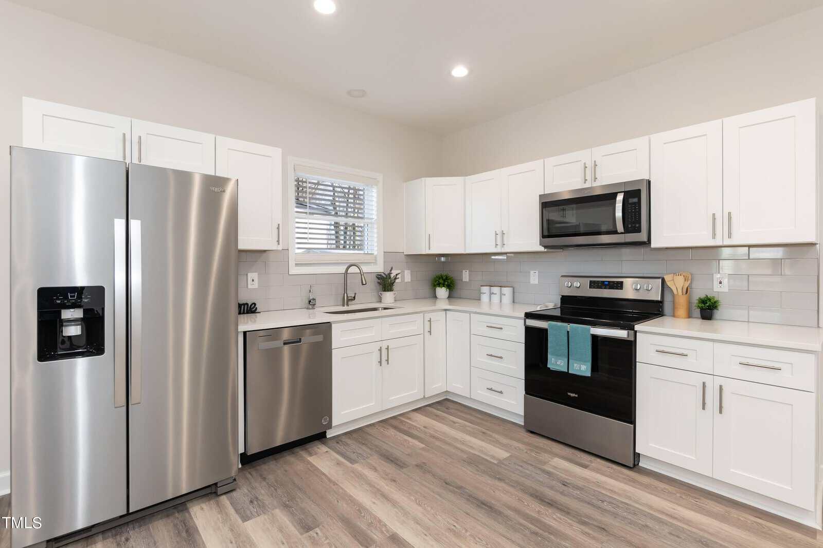 Modern kitchen with stainless steel appliances, including a refrigerator, dishwasher, and oven with microwave. White cabinets, gray tile backsplash, and wood flooring. A window above the sink and decor items on the counter.
