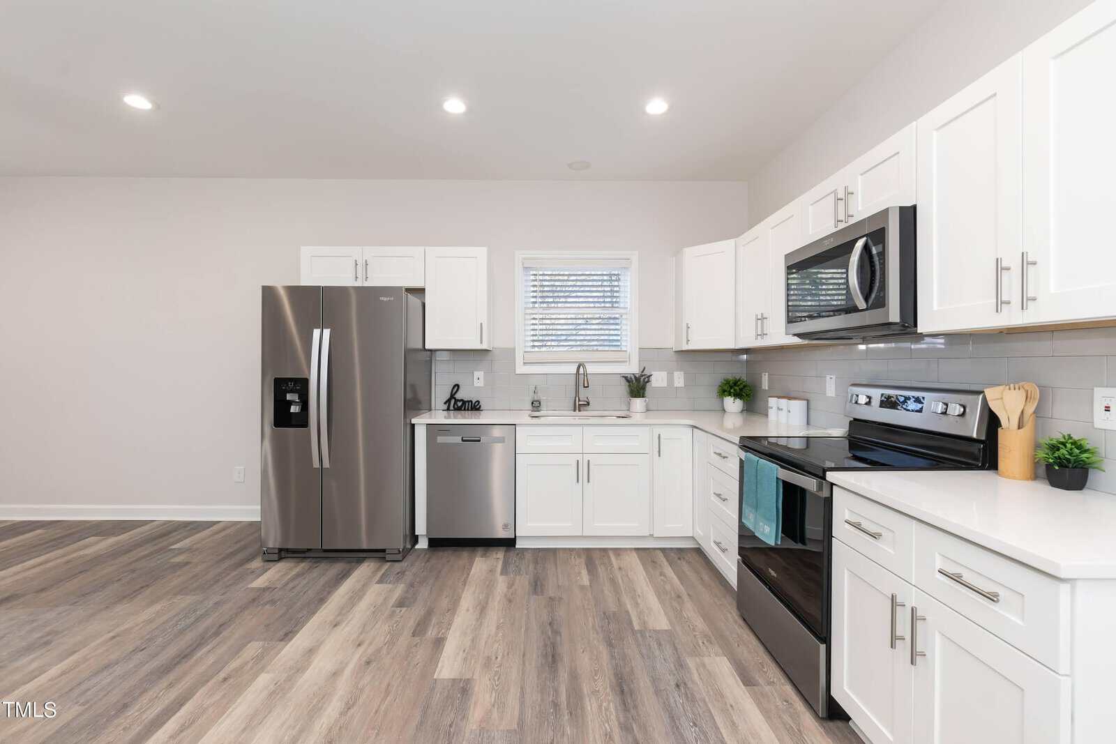Modern kitchen with white cabinets, stainless steel appliances including a refrigerator, oven, and dishwasher. Light wood flooring complements the clean design. Small plants decorate the countertop for a touch of greenery.
