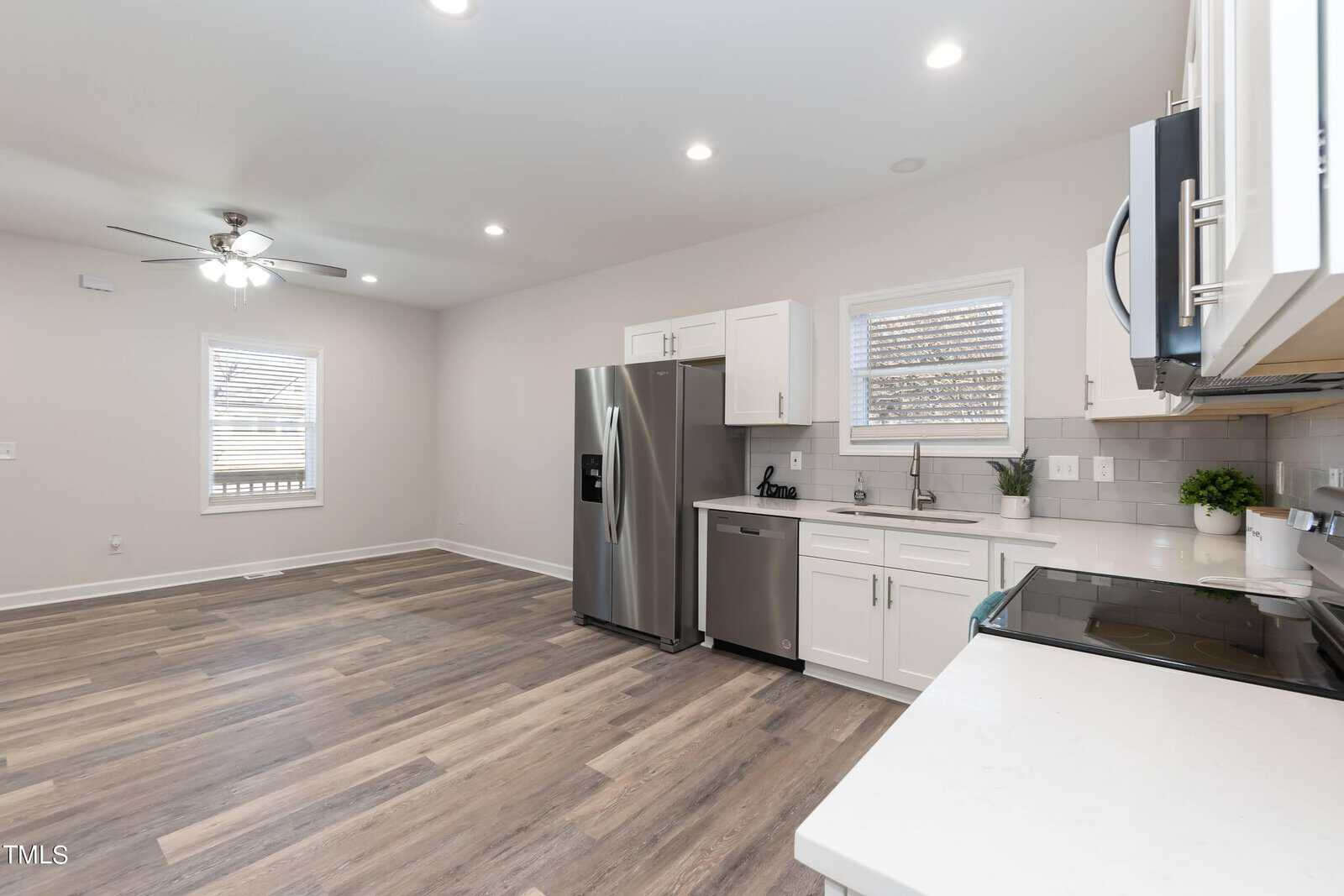 Bright, modern kitchen with stainless steel appliances, white cabinets, and wood flooring. Open layout includes a ceiling fan and recessed lighting, with a window above the sink and a spacious area adjacent to the kitchen.