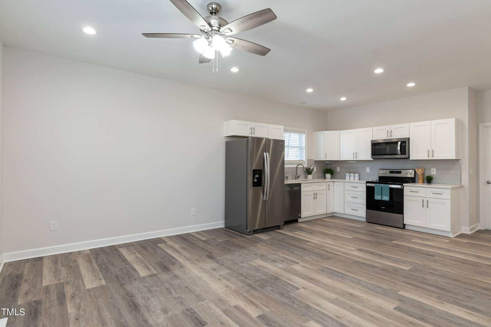 A modern kitchen with white cabinets, stainless steel appliances including a fridge and oven. It features wooden flooring, a ceiling fan with lights, and an uncluttered countertop near a window. The room is spacious and well-lit.