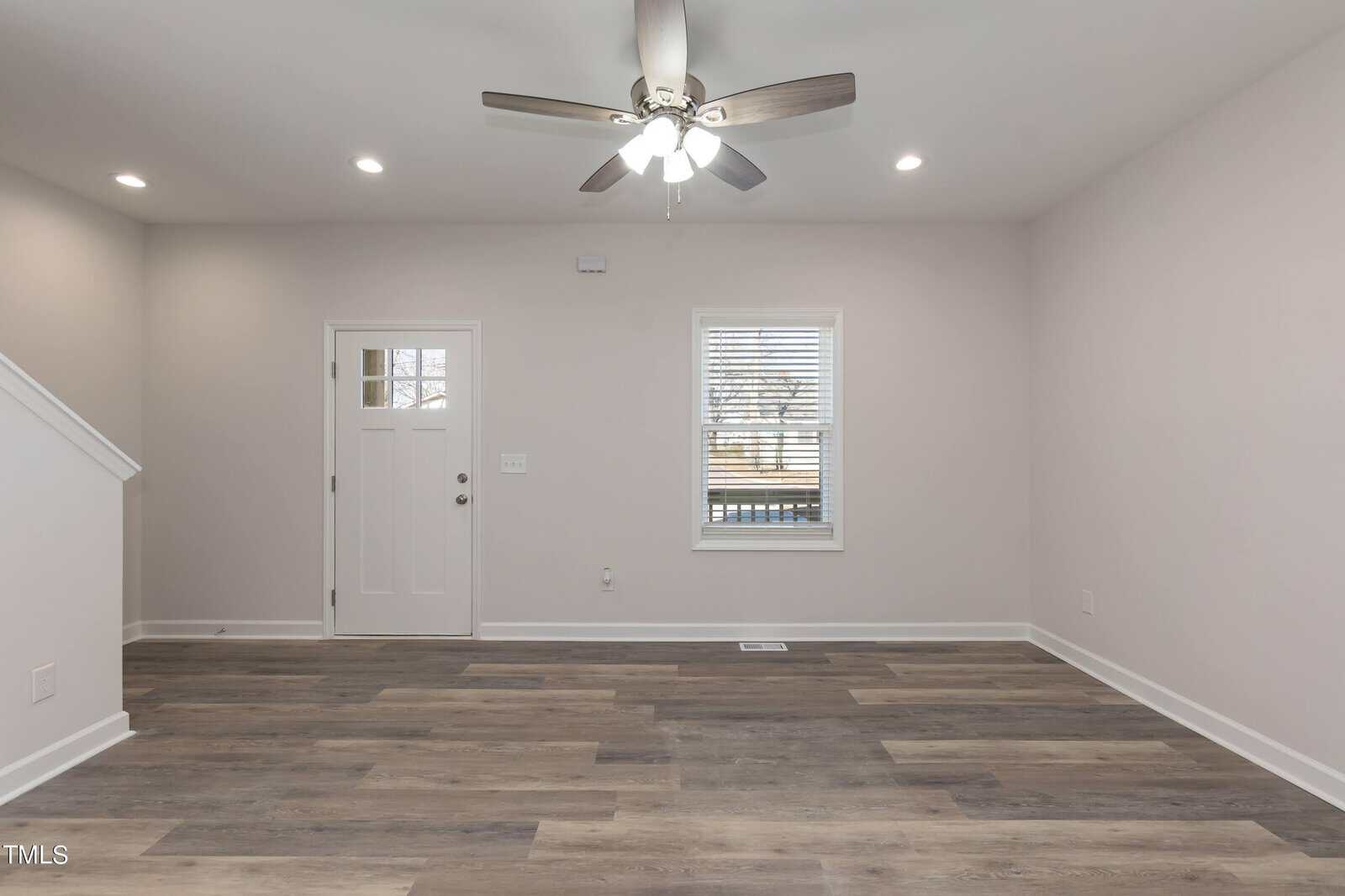 An empty living room with light gray walls and wood-look flooring. A ceiling fan with lights is mounted on the ceiling. The room features a door and a window with blinds, and there's a staircase on the left side.