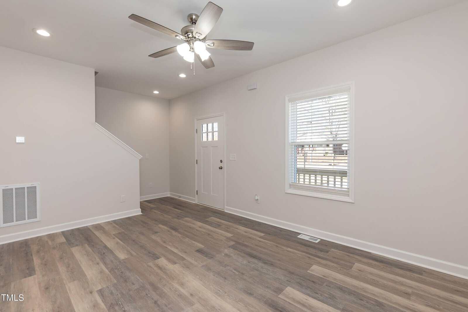 A bright, empty room with light gray walls and wood flooring. A ceiling fan with lights hangs from the ceiling. A window with blinds is on the right, and a door with a small window is on the wall opposite the stairs.