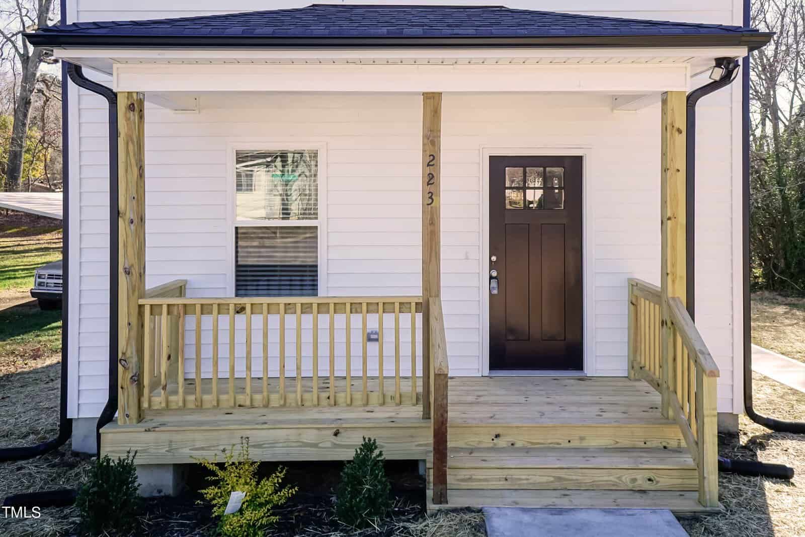Front view of a house with a small wooden porch, stairs, and railing. The door is dark-colored, and the exterior walls are white. A window with closed blinds is to the left of the door. Small plants are in front of the porch.