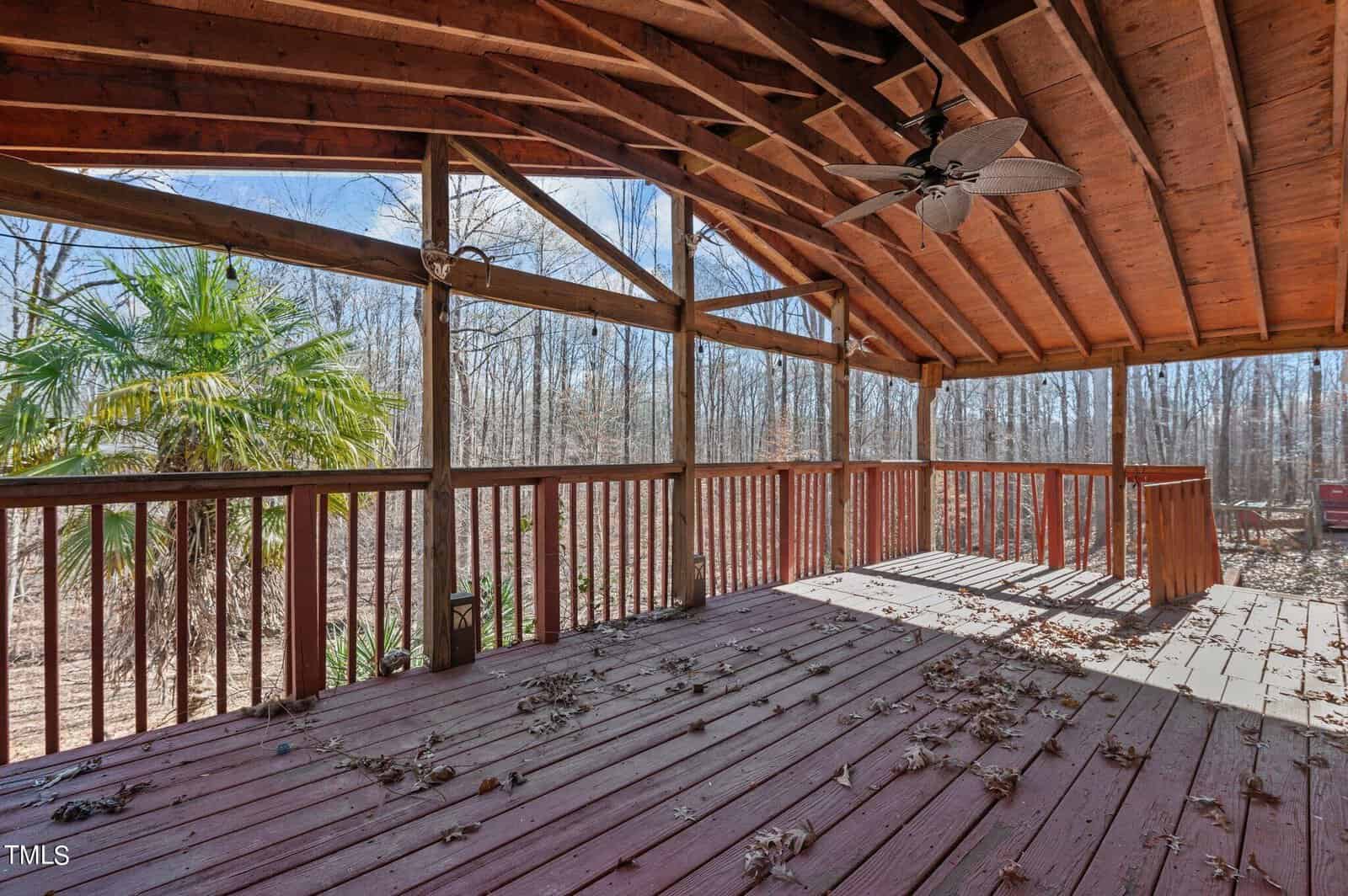 A wooden deck with a high, angled roof and ceiling fan overlooks a wooded area. The deck is surrounded by railings, with fallen leaves scattered on the wooden floor. A palm tree stands on the left side, enhancing a rustic, outdoor atmosphere.